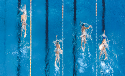 Fotografia com vista aérea de 4 nadadores em uma piscina, cada um em uma raia, estando um mais a frente que os demais.