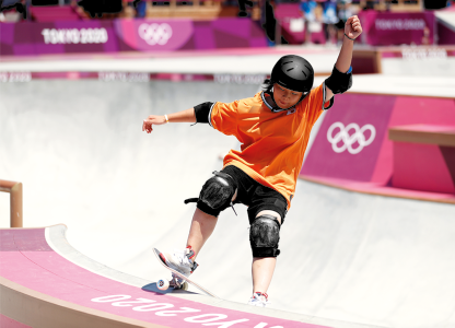 Fotografia de um menino em cima de um skate fazendo manobras em uma pista. Ele usa capacete e joelheiras, e em uma das estruturas está o símbolo das olimpíadas.