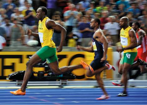 Fotografia de 4 corredores em uma pista de corrida, com um atleta na frente dos demais. Ele usa regata amarela e shorts verdes. Ao fundo, há uma plateia em uma arquibancada.