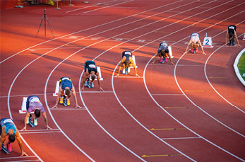 Fotografia de vários homens em posição de largada, abaixados, em uma pista de corrida curva. Eles estão posicionados da seguinte forma: quanto mais na circunferência de fora, mais a frente ele está.