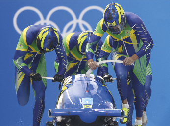 Fotografia de quatro pessoas uniformizadas, usando capacete e macacão nas cores azul, amarelo e verde. Eles estão empurrando um trenó juntos, e ao fundo há o símbolo das olimpíadas.