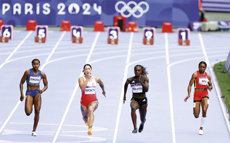 Fotografia de quatro mulheres com uniformes diferentes correndo em uma pista, cada uma em sua raia, e ao fundo está escrito Paris 2024 e o símbolo das Olimpíadas.