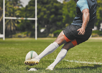 Fotografia de uma pessoa com roupa esportiva, camiseta, shorts, meião e chuteira. Ela está em um gramado chutando uma bola de formato oval, em direção a uma trave ao fundo que tem formato de H.