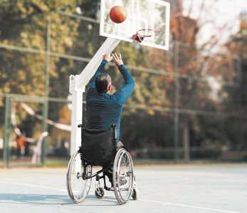 Fotografia de um homem em uma cadeira de rodas em uma quadra arremessando uma bola na direção de uma cesta de basquete.