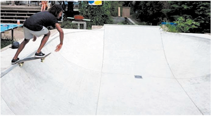 Fotografia de uma pessoa fazendo manobras com um skate em uma pista. Ela é vista de costas sobre o skate, com um pé em cada extremidade, sendo que a extremidade direita está para fora da rampa.