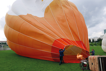 Fotografia de um balão de ar quente, semicheio, deitado na grama com duas pessoas o segurando, enquanto há uma chama no sentido da área interna.