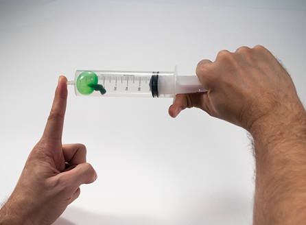 Fotografia de duas mãos segurando uma seringa com um balão verde dentro. A mão direita está puxando o êmbolo e a mão esquerda está tampando a ponta da seringa, com isso o balão está com volume maior comparado ao anterior.