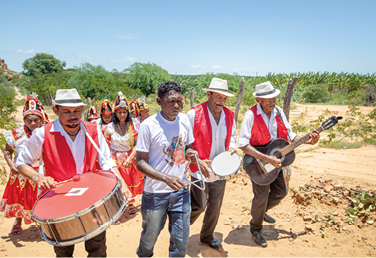 Fotografia de um grupo de pessoas andando em uma estrada de terra com vegetação ao fundo. Há quatro homens tocando instrumentos musicais na frente, como tambor, triângulo, pandeiro e violão; e atrás há algumas mulheres dançando. Os homens usam camiseta branca, na qual três também estão de chapéu e usam coletes vermelhos. As mulheres usam um chapéu adornado na cabeça e vestidos com as cores branco e vermelho.