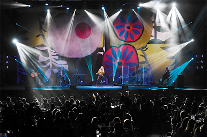 Fotografia de um show com uma banda musical em um palco, na qual uma pessoa está no centro, cantando ao microfone. No palco há muitas luzes e ao fundo há desenhos coloridos. Em frente ao palco há muitas pessoas na plateia que está escura.