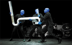 Fotografia de três pessoas vestidas de preto e com rosto e mãos pintados de azul. Elas estão segurando uma estrutura feita com canos brancos interligados. Ao fundo há vários objetos escuros.