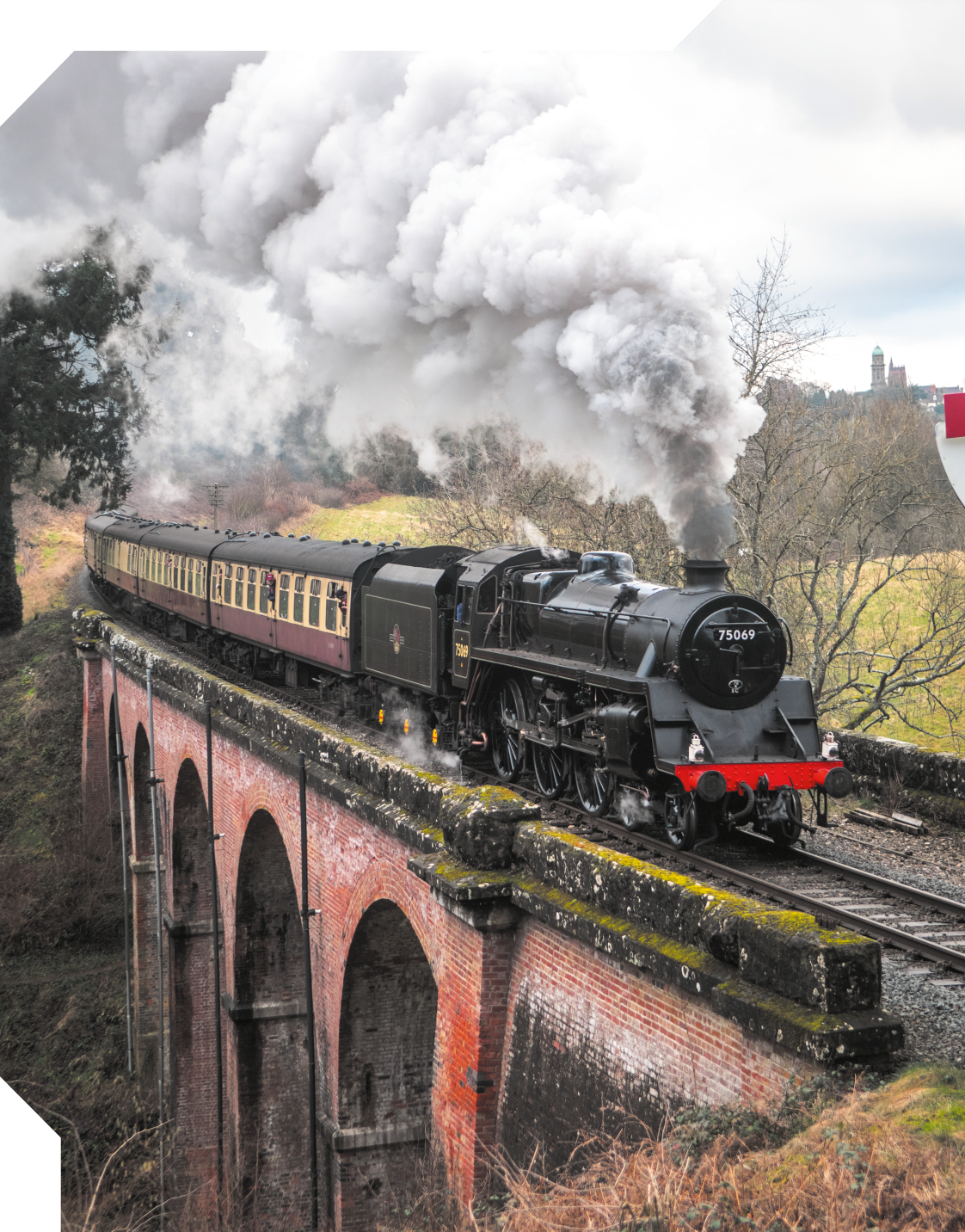 Fotografia de uma locomotiva com alguns vagões passando por trilhos no alto de uma ponte. Na parte frontal há fumaça saindo de um tubo superior. Ao redor há árvores e gramados.