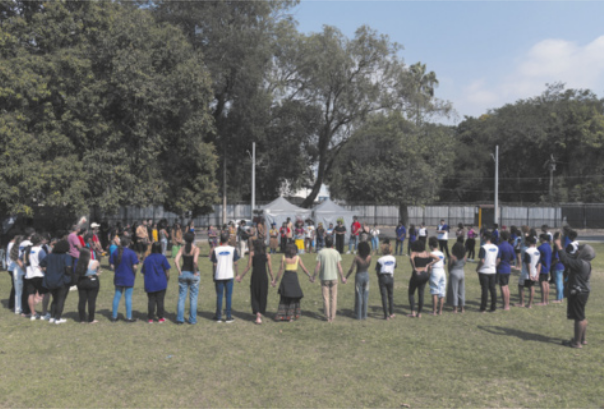 Foto de pessoas reunidas em um espaço aberto. Elas estão em roda e de mãos dadas.