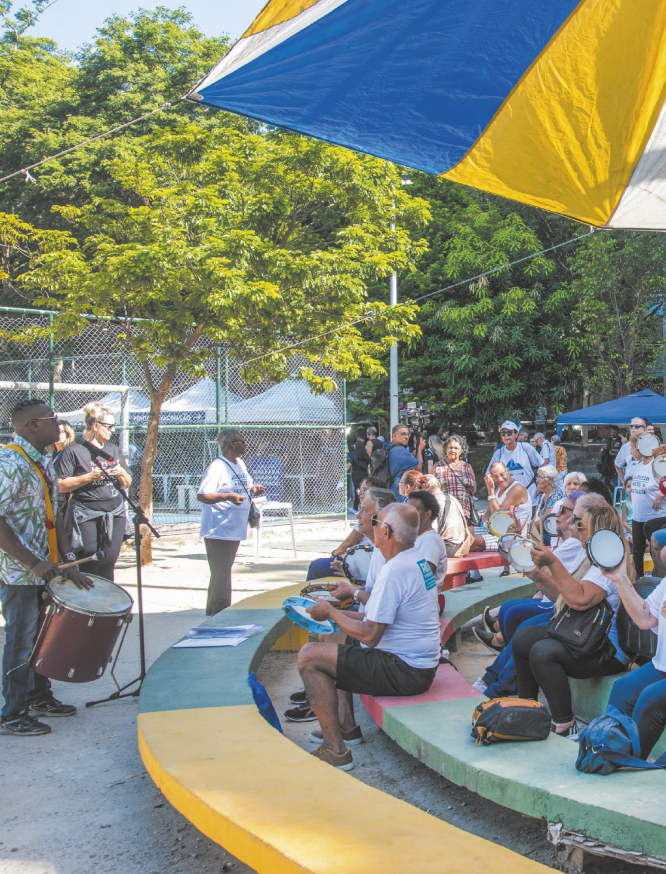 Foto de pessoas tocando tambor, caixa e pandeiros. Enquanto isso, os demais assistem.