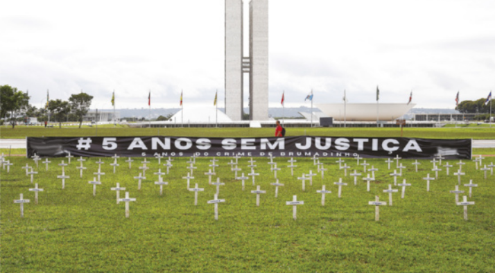 Foto de protesto. Neste protesto, foram instaladas muitas cruzes e uma faixa, onde está escrito 'Hashtag 5 anos sem justiça'. Ao fundo, encontra-se um edifício formado por uma estrutura horizontal, sobre a qual se encontram duas semiesferas.