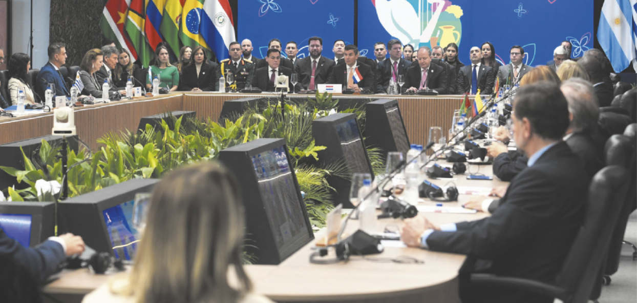 Foto de pessoas reunidas em um salão. Elas se comunicam usando microfones e utilizam computadores. Também se destacam as bandeiras de diferentes países.