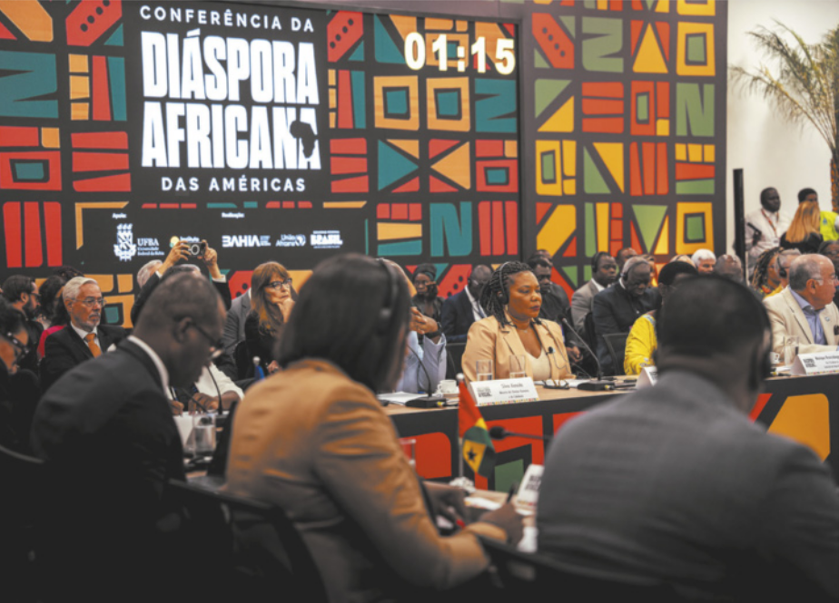 Foto de pessoas reunidas em um salão. Elas se comunicam usando microfones e utilizam computadores. Também se destaca um banner, onde está escrito 'Conferência da Diáspora Africana das Américas'.