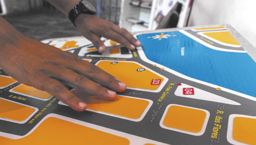 Foto de pessoas manuseando um mapa tátil. Neste mapa, apresentam-se ruas e quarteirões.