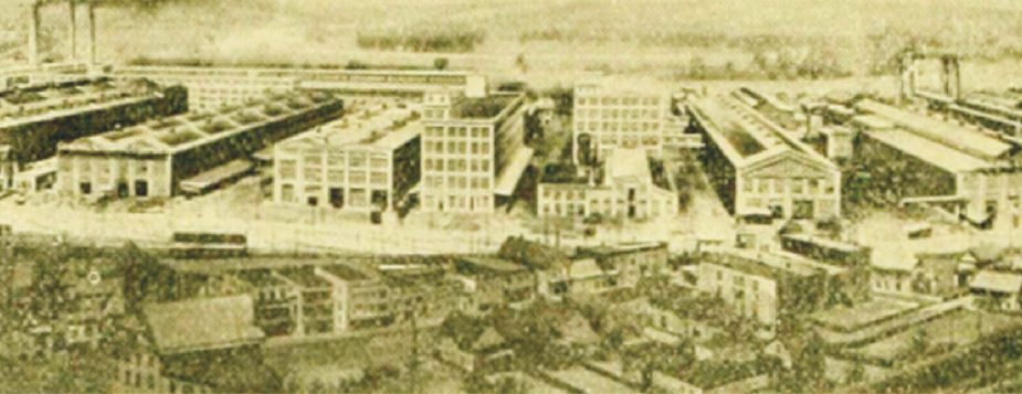 Foto de fábrica formada por grandes galpões e edifícios com muitos andares. Ao fundo, destacam-se chaminés.
