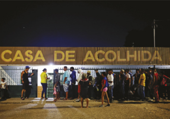 Foto de edifício com fila na parte exterior. Na fachada, está escrito 'Casa de acolhida'.
