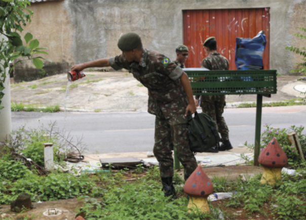 Foto de homens usando uniforme militar. Eles retiram a água que está acumulada em embalagens e outros locais.