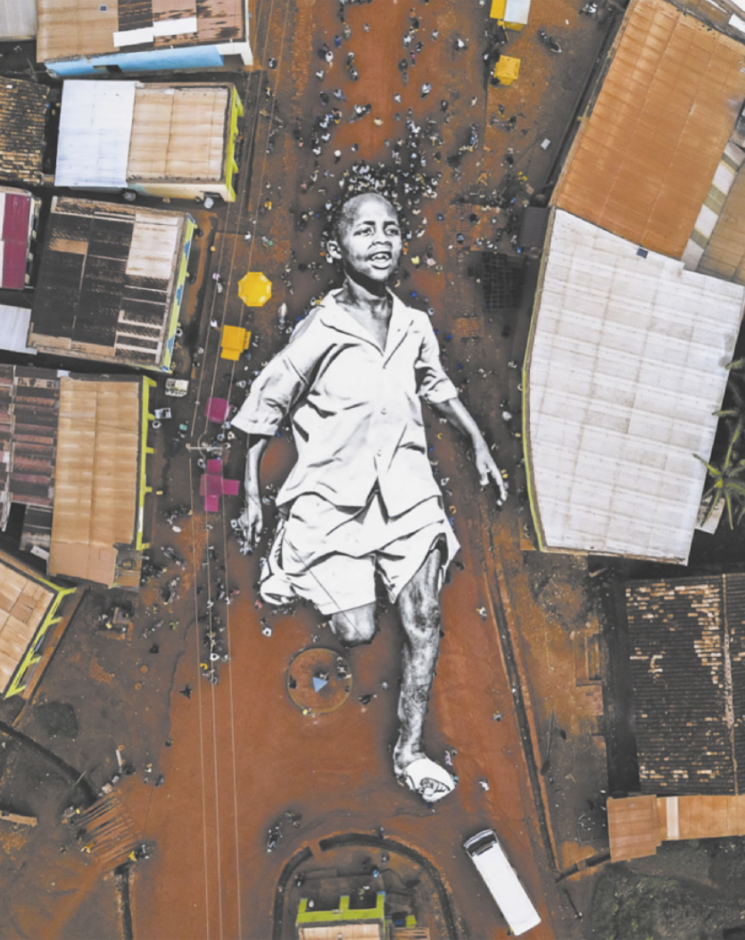 Foto aérea, vista de cima, de uma rua larga de terra. Há uma de obra de arte composta por grandes fotografias coladas no chão, formando a imagem de um menino correndo. Ele está sorrindo, tem cabelos raspados e usa camisa e bermudas claras. Há pessoas circulando e um ônibus, que tem aproximadamente o tamanho do antebraço do garoto da imagem.