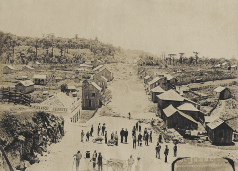 Foto de pequenas moradias, feitas de madeira. Essas moradias estão ao redor de uma rua, onde as pessoas caminham.