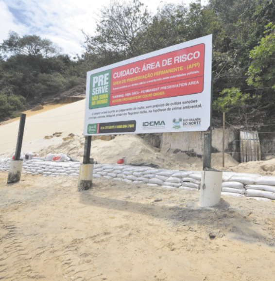 Foto de placa instalada na areia da praia. Na placa, está escrito 'Cuidado: área de risco. Área de preservação permanente. Preserve, não suba o morro'.