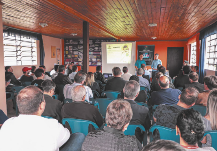 Foto de pessoas reunidas em uma sala. Alguns estão a frente e falam para os demais.