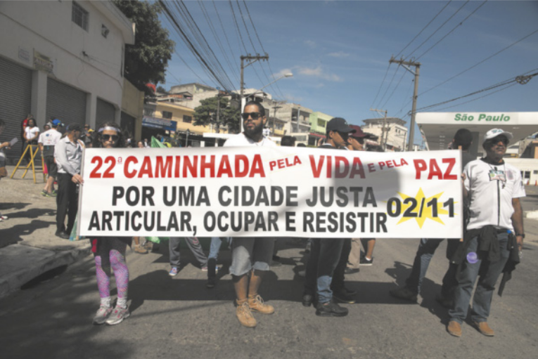 Foto de pessoas em manifestação. Elas seguram uma faixa, onde está escrito 'Vigésima segunda caminhada pela vida, pela paz. Por uma cidade justa. Articular, ocupar e resistir'.