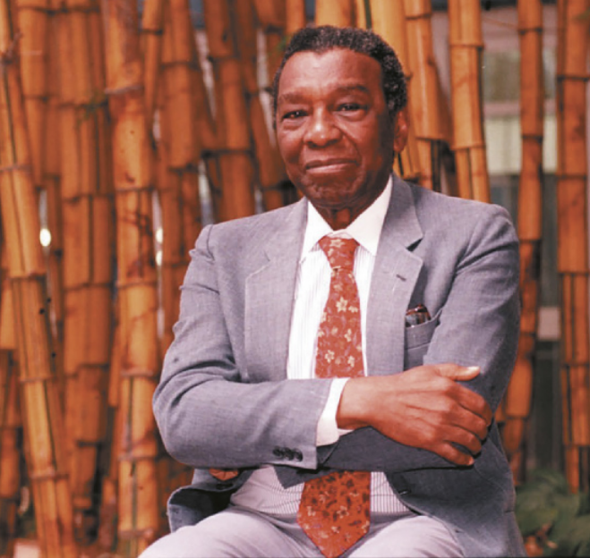 Foto de Milton Santos. Ele é um homem negro, possui cabelos curtos e usa terno.