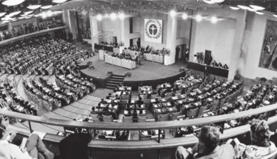 Foto de muitas pessoas reunidas em um auditório. No palco, há uma mesa onde estão algumas pessoas.