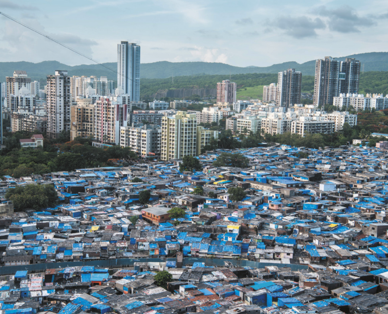 Foto de pequenas moradias, construídas muito próximas umas das outras. Ao fundo, destacam-se prédios altos.