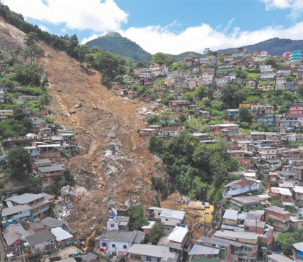 Foto de deslizamento de terra em área inclinada. É possível identificar escombros de construções por onde a terra deslizou. Também há terra acumulada ao lado das casas instaladas em locais mais baixos.