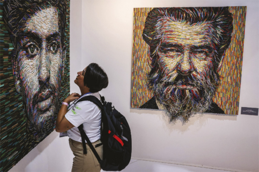 Foto de mulher observando quadros. Esses quadros estão expostos na parede e apresentam o rosto de dois homens.