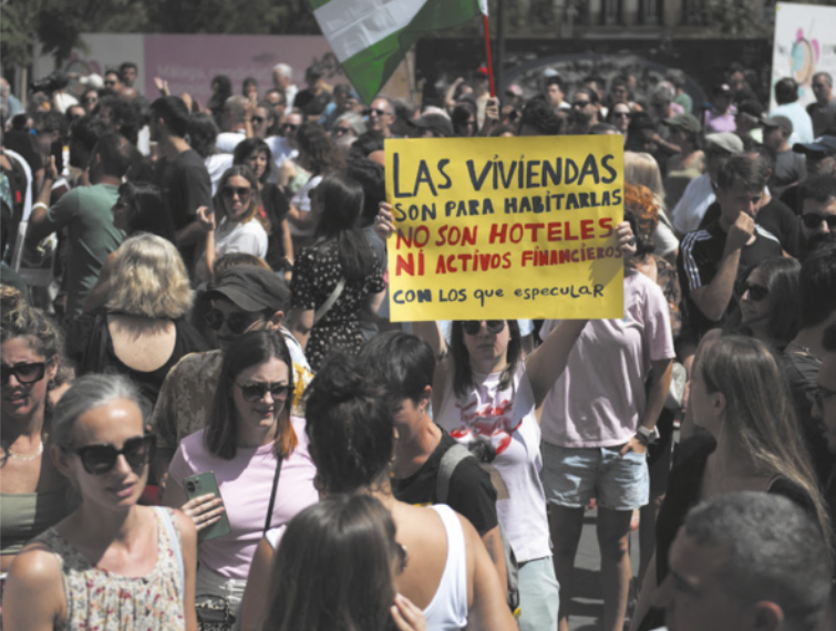 Foto de pessoas em manifestação, destaca-se uma mulher segurando um cartaz com dizeres em espanhol.