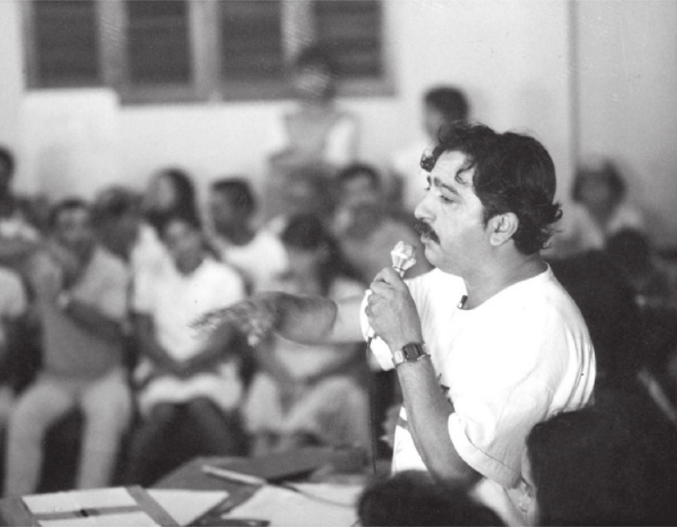 Foto de Chico Mendes. Ele é um homem branco, usa bigode e está dando uma palestra.