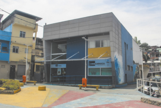 Foto de praça circular com bancos e jardins. À frente, há uma construção de dois andares, além de diversas casas ao redor.