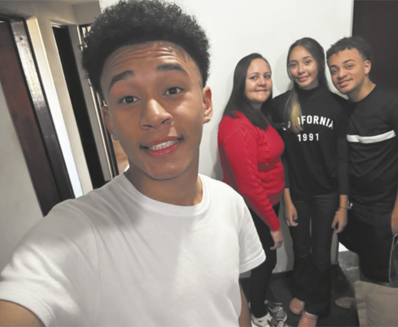 Foto de menino. Ele tira uma fotografia de si e, ao fundo, destaca-se uma mulher, um jovem e uma jovem.