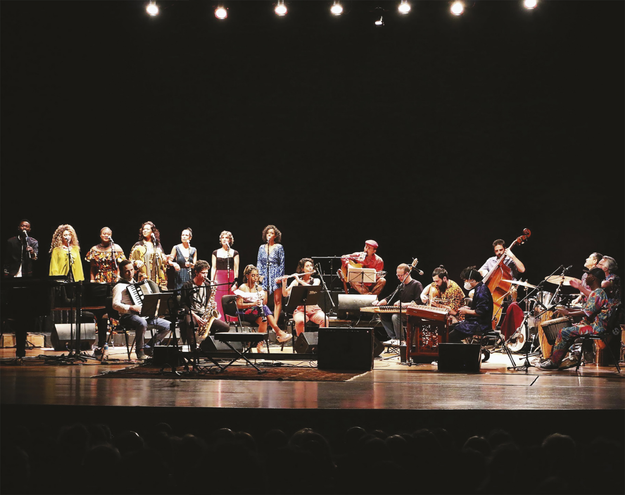 Foto de pessoas em apresentação, elas tocam diferentes instrumentos, como flauta, clarinete, violoncelo, tambor e bateria.