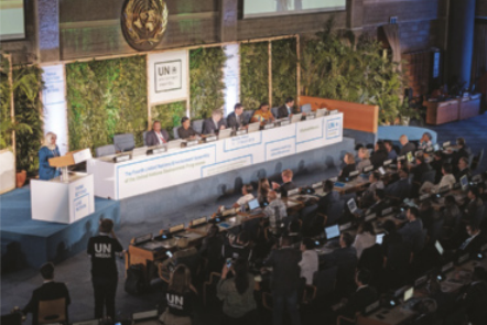 Foto de seis pessoas sentadas ao redor de uma mesa, em um palco. Neste palco, também se destaca uma mulher em pé, discursando.