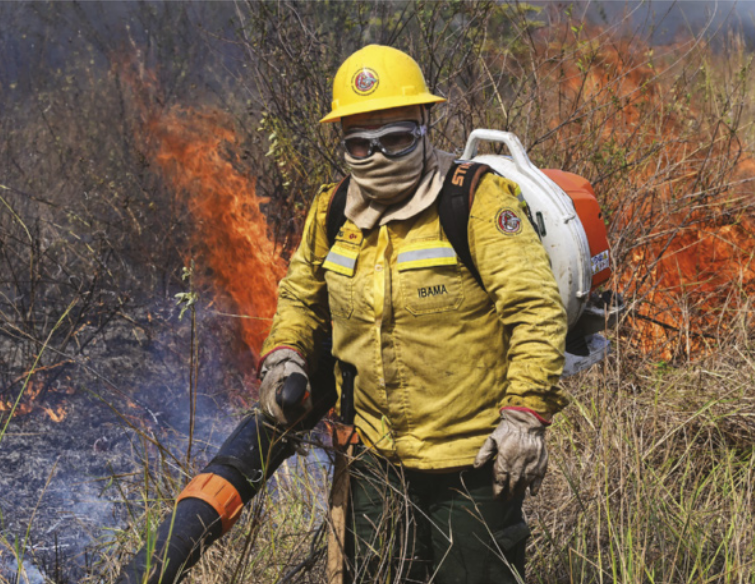 Foto de homem combatendo um incêndio. Ele usa capacete, roupa especial, luva, protetor para os olhos e um tecido, que cobre o nariz e a boca.