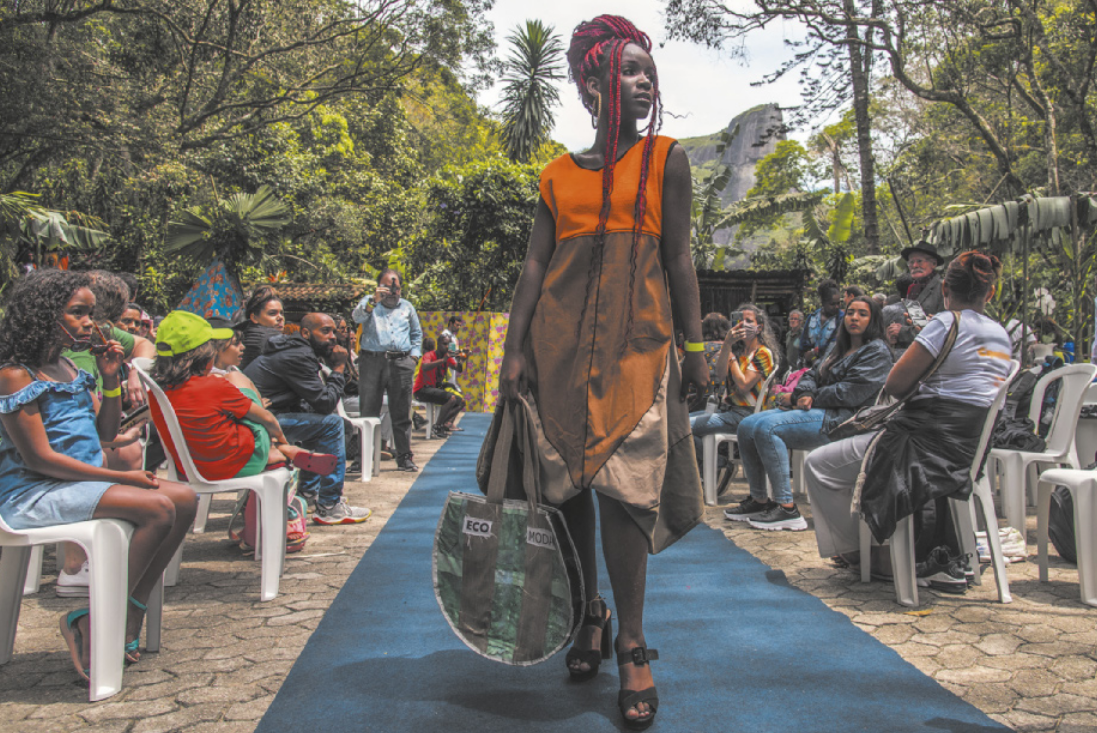 Foto de mulher negra desfilando. Ela usa um vestido com diferentes tecidos, uma bolsa e tranças vermelhas.