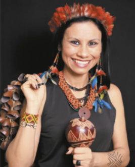 Foto de 'Márcia Wayna Kambeba'. Ela é uma mulher indígena, usa cocar, segura um chocalho e está sorrindo.