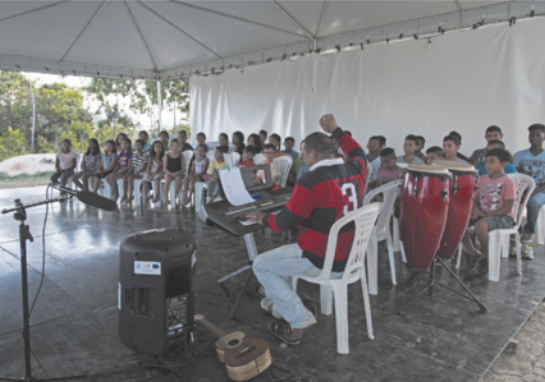 Foto de crianças e jovens reunidos. Eles estão sentados e cantando. Enquanto isso, um homem toca teclado.