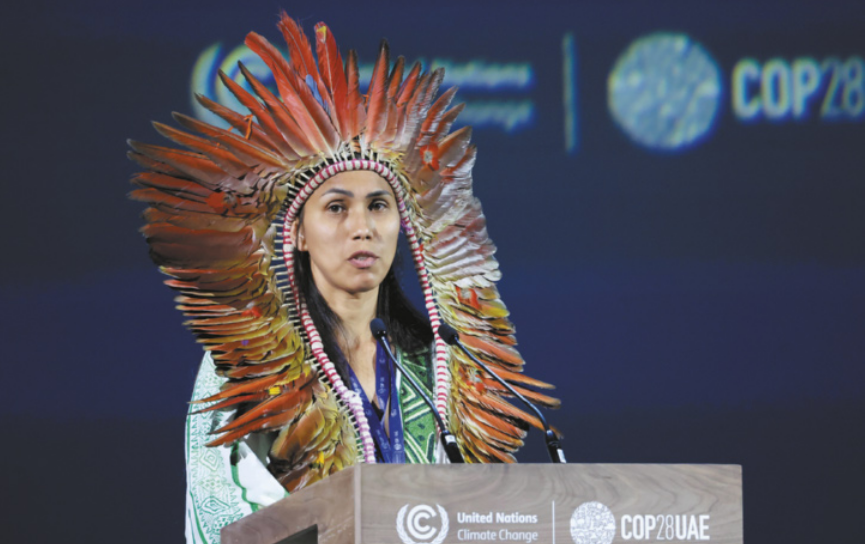 Foto de Isabel Gakran. Ela é uma mulher indígena, está discursando e usa um cocar.