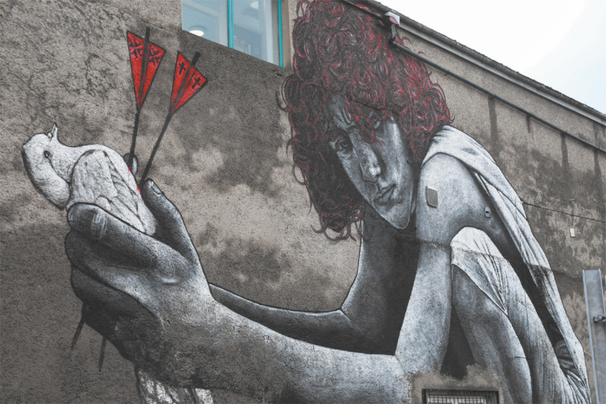 Mural que apresenta uma pessoa agachada e segurando uma pomba branca. Essa pomba foi atingida por duas flechas, uma possui o símbolo dos católicos e outra o símbolo dos protestantes.