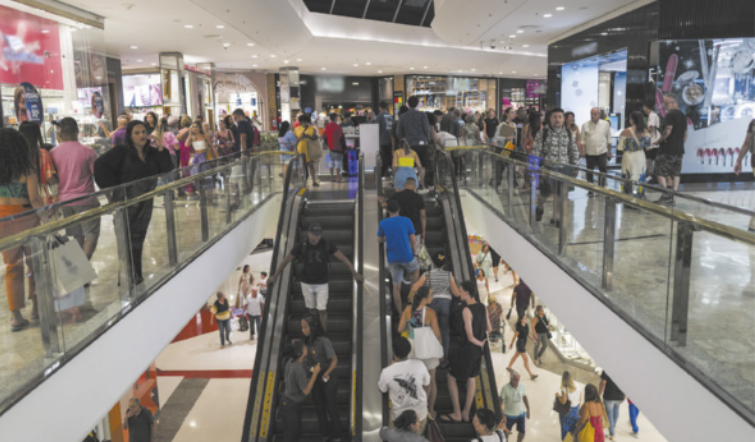 Foto de pessoas no shopping. Elas caminham pelo corredor e usam a escada rolante.