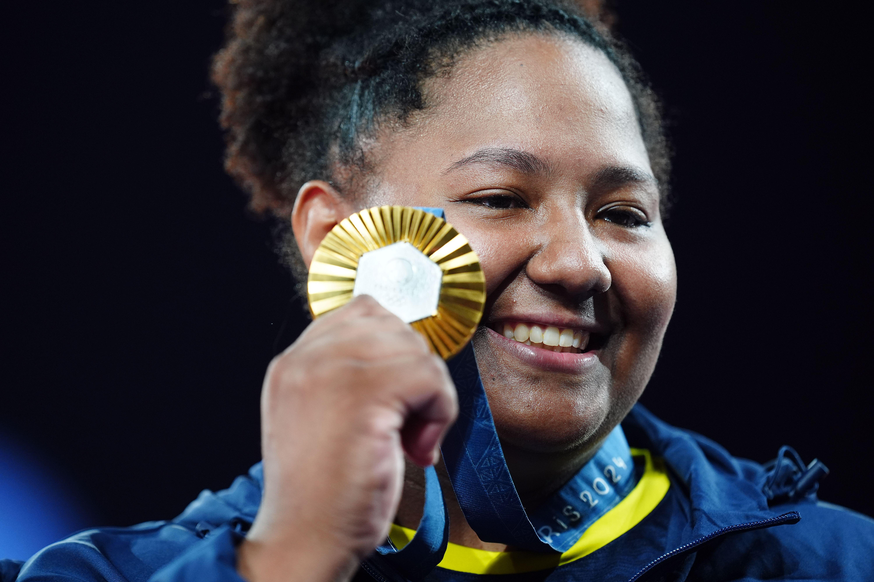 Fotografia de busto de uma mulher sorrindo enquanto segura uma medalha de ouro ao lado do rosto. Ela é negra, está com cabelos presos e usa roupa nas cores azul e amarelo.