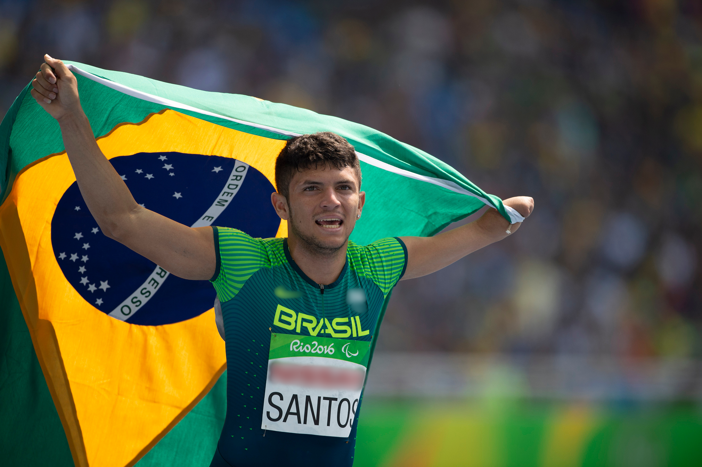 Fotografia de um homem segurando uma bandeira do Brasil por trás do corpo. Ele é branco, não tem parte de um dos braços e usa uma camiseta em tons de verde com a escrita Brasil, Rio 2016.