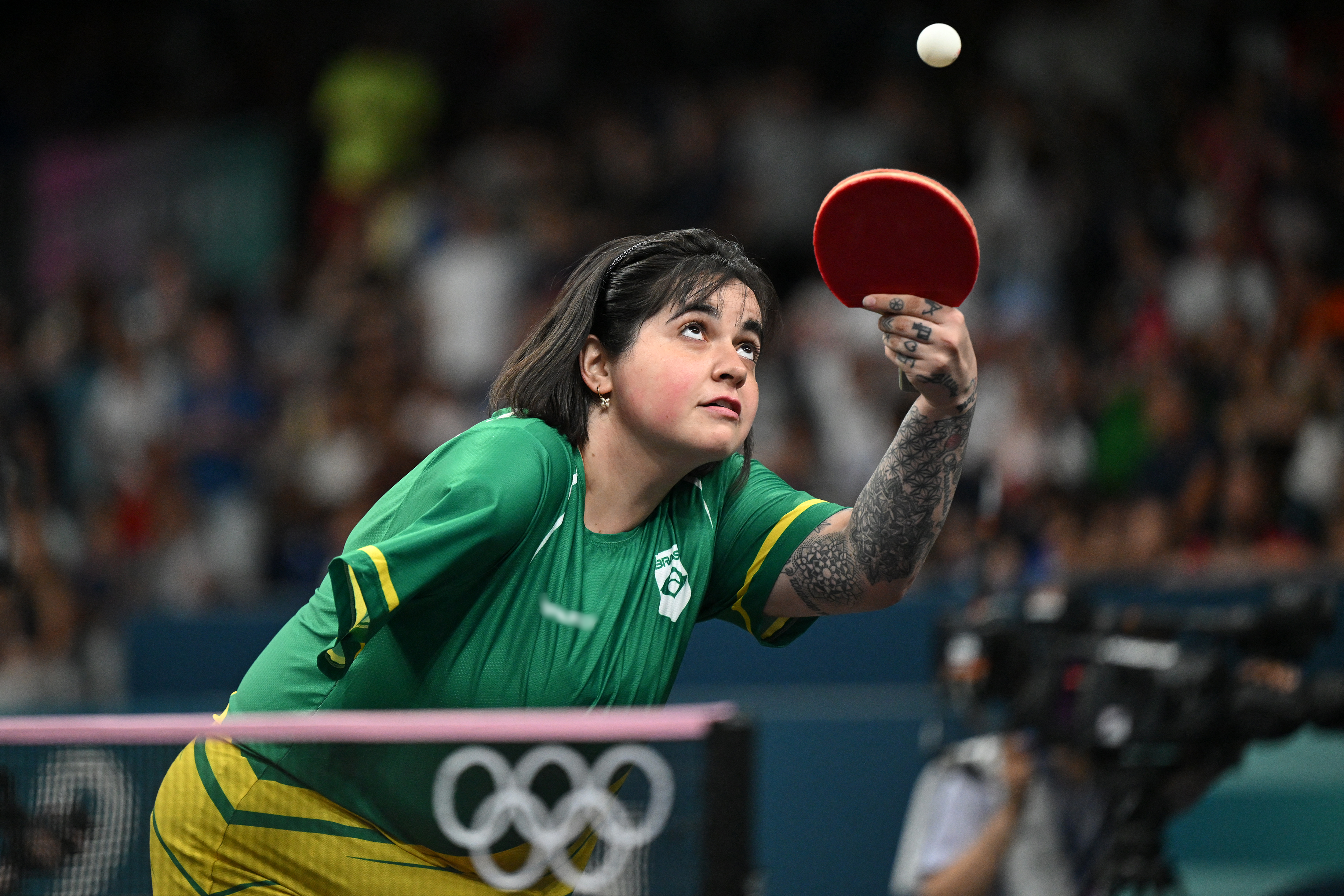 Fotografia de uma mulher durante uma partida de tênis de mesa. Ela é branca, tem várias tatuagens no braço esquerdo e não tem o braço direito. Usa uma camiseta nas cores verde e amarelo enquanto olha para a bolinha que está no ar, acima da raquete, que ela segura com a mão esquerda.
